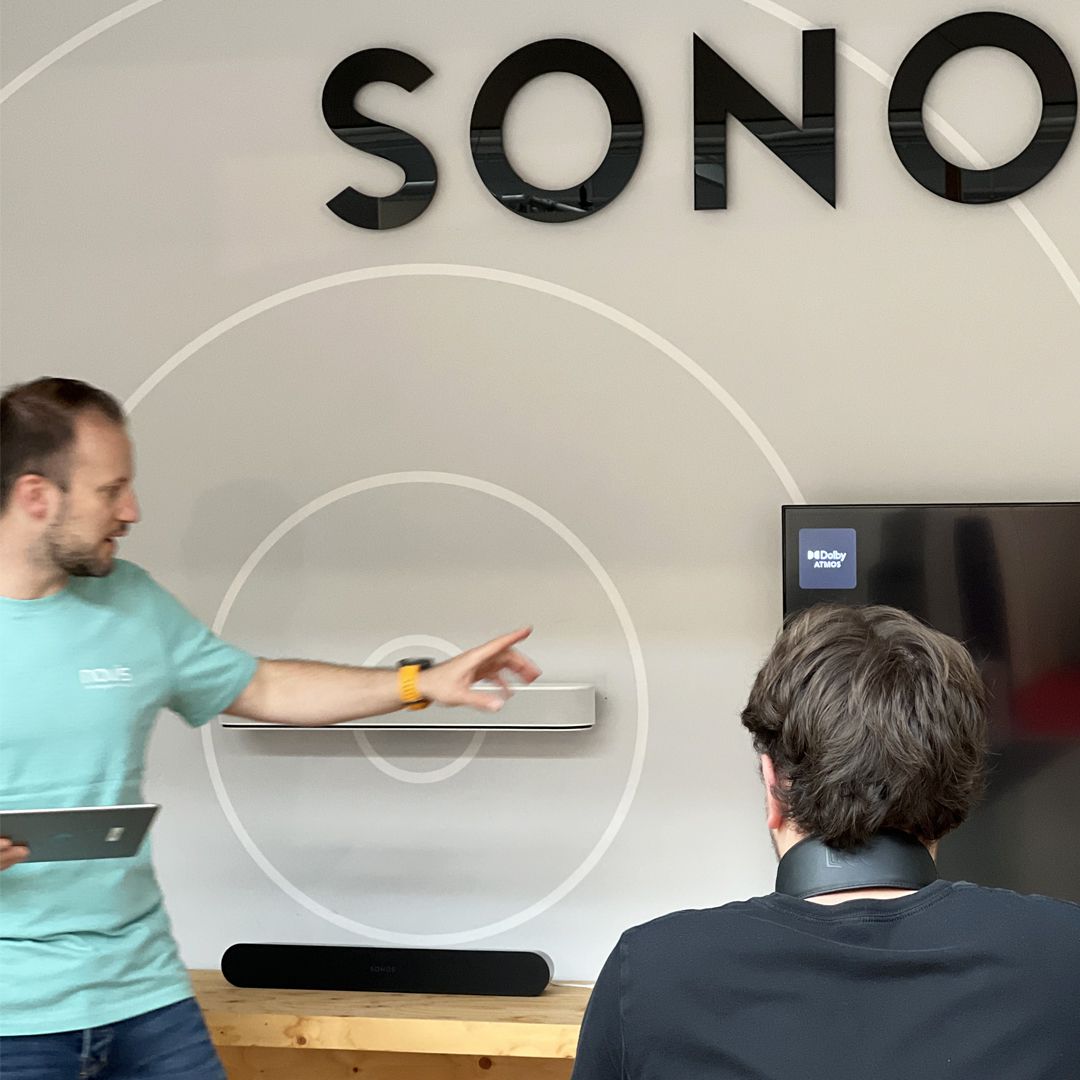 Eine männliche Person führt eine Demo durch von Sonos Lautsprechern. Eine andere Person sieht dabei zu