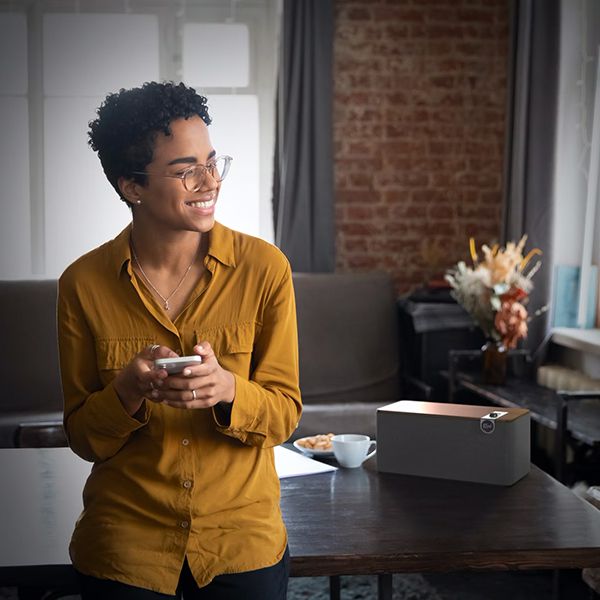 Lifestyle Bild: Eine junge Frau im senfgelbem Hemd mit dem Handy in der Hand steuert ihr Premium Bluetooth Speaker von Klipsch