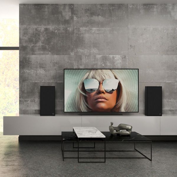 Black speakers on a white sideboard. In the middle of the sideboard is a TV with a woman and sunglasses. There is also a sofa and a side table in the room.