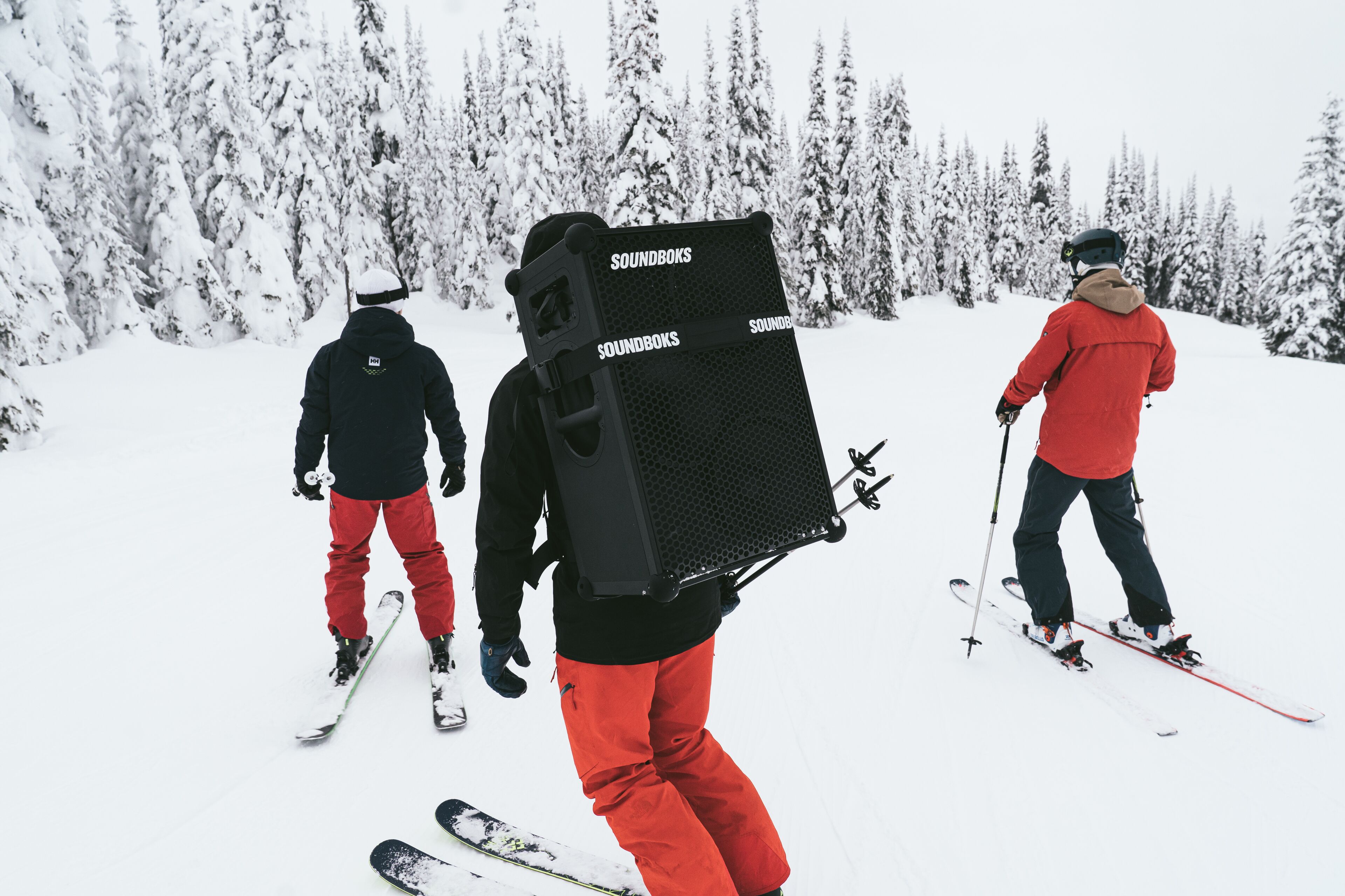 3 Personen am Skifahren im Tiefschnee und ein Skifahrer hat einen Soundbox Lautsprecher am Rücken