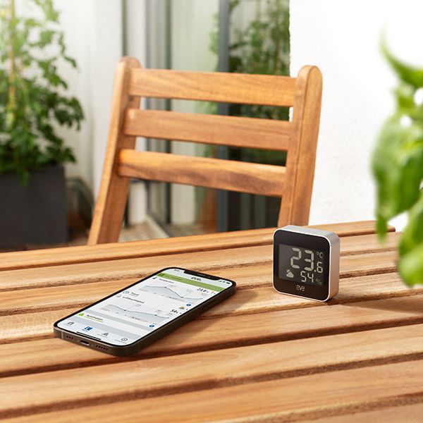 Auf einem hölzernen Gartentisch steht das Eve Weather und daneben ein Handy, worauf die App des Eve Weather dargestellt wird